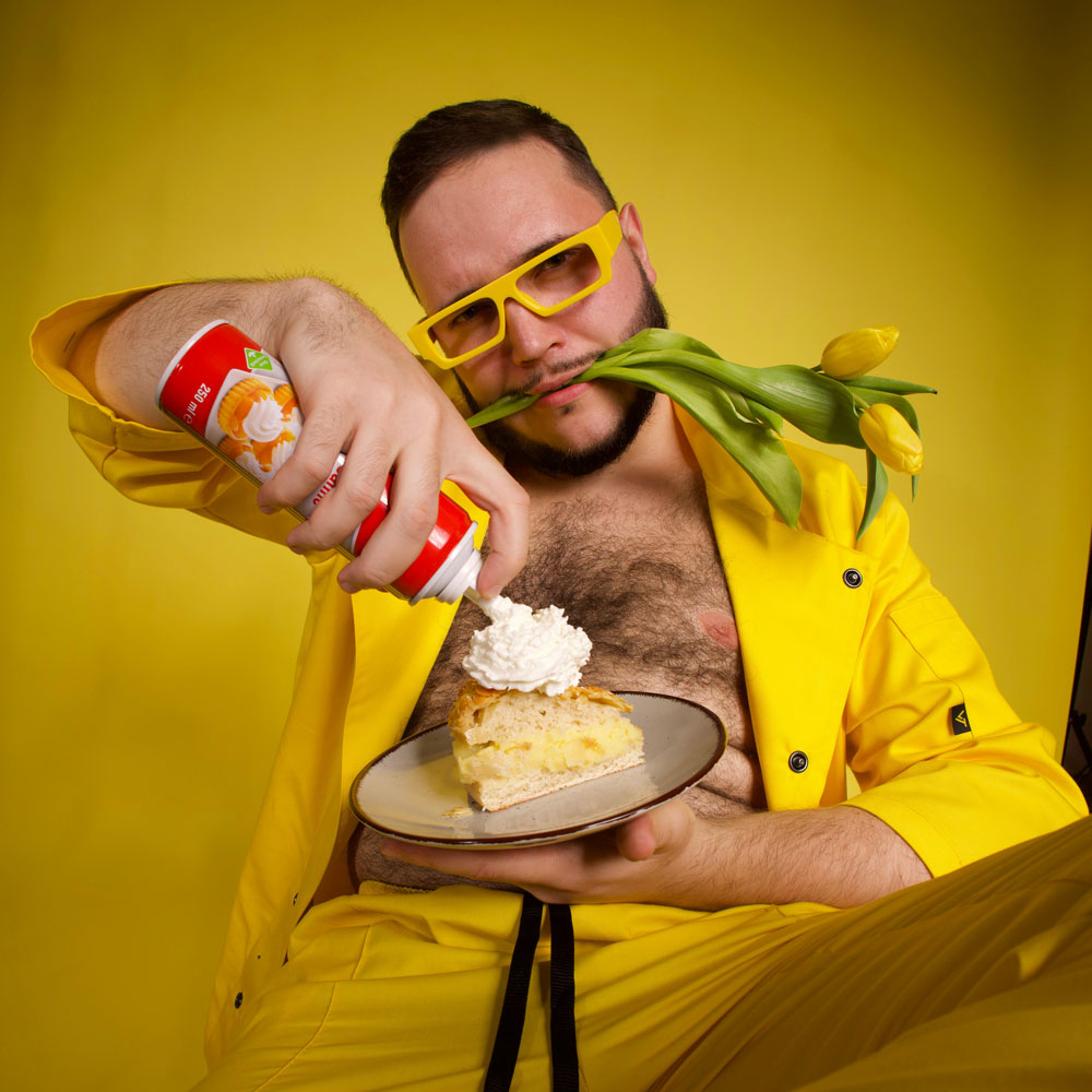 A man wearing yellow sunglasses and an open shirt sprays cream onto a piece of cake.